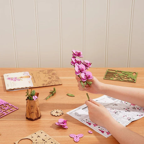 Person assembling Purple Moth Orchid DIY Wooden Flower on a wooden table with craft materials.