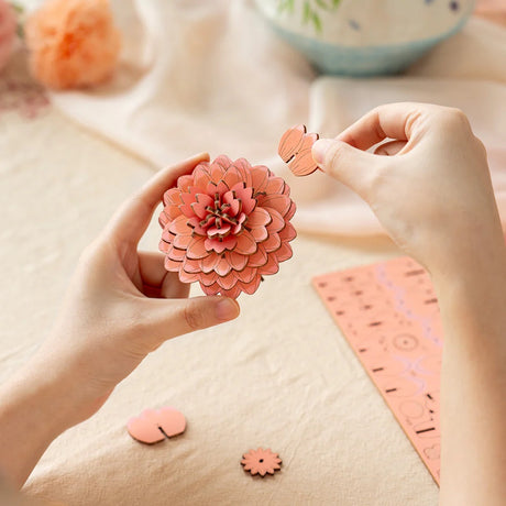 Person assembling a Pink Dahlia DIY Wooden Flower 3D Puzzle with craft materials on a table.