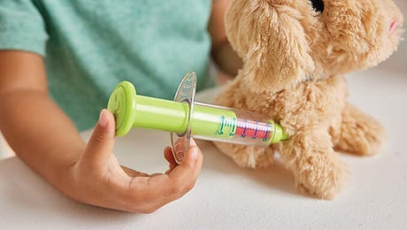 Person holding a green syringe-like toy next to a stuffed dog on a light surface.