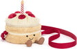 A plush cake-shaped crossbody bag with a cream-colored frosting top, red cherries, a red-and-white striped candle, and a red strap, shown on a white background.