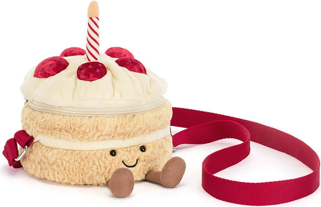 A plush cake-shaped crossbody bag with a cream-colored frosting top, red cherries, a red-and-white striped candle, and a red strap, shown on a white background.