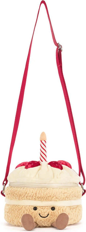 A plush Amuseables Birthday Cake Bag designed like a cute cream-colored cake with a red ribbon and candle, featuring a smiling face and a red shoulder strap on a white background.