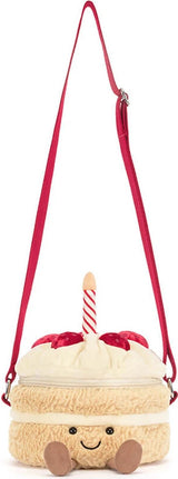 A plush Amuseables Birthday Cake Bag designed like a cute cream-colored cake with a red ribbon and candle, featuring a smiling face and a red shoulder strap on a white background.
