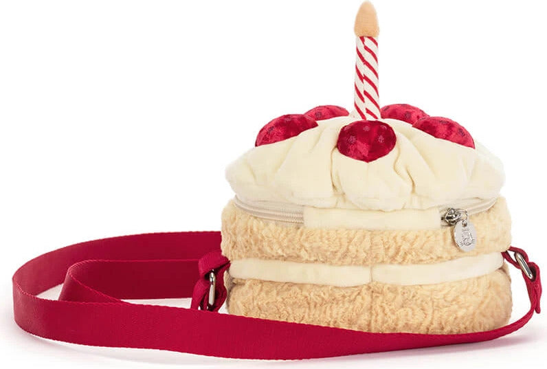 A plush Amuseables Birthday Cake Bag with cream-colored faux frosting, red cherry details, a red waist strap, and a single red-and-white striped candle on top, shown on a white background.