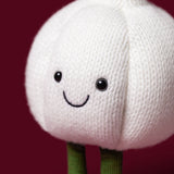 A close-up of a Amuseables Garlic character toy with a white textured body, embroidered smiling face, and green stem-like legs against a dark red background.
