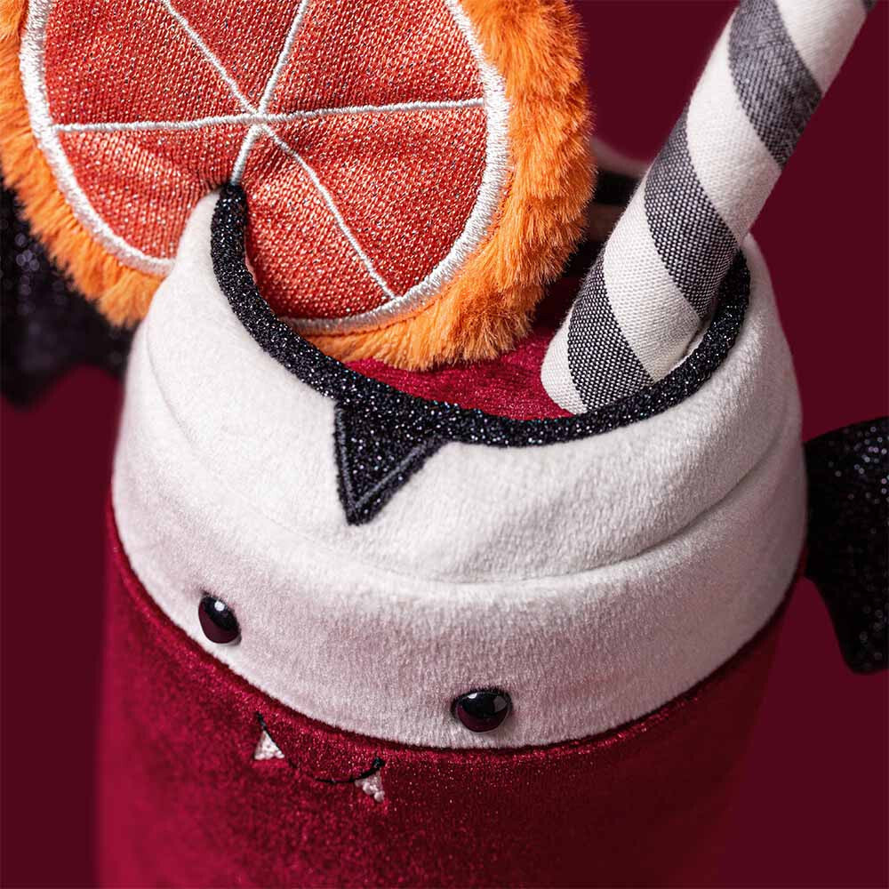 A close-up of a plush Amuseables Vampop Drink, featuring a red velvet-like body, white plush whipped cream top, black glitter trim, a striped gray-and-white straw, and an orange slice accent on a dark red background.