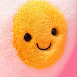 Close-up of the smiling face on the soft yellow center of the Amuseables Heart Boiled Egg plush toy with a pink background.
