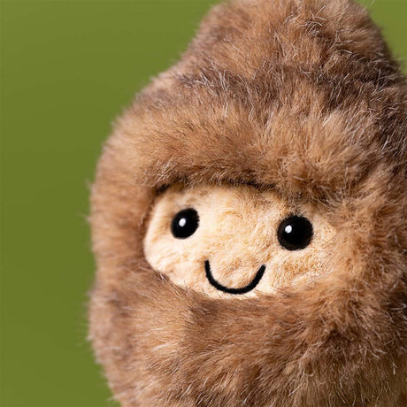 Close-up of a plush Amuseables Peanut Yeti Outfit toy with a brown fuzzy exterior and beige face on a green background