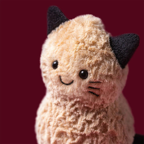 A close-up of a plush Amuseables Peanut Cat Outfit toy with fluffy cream-colored fur, black ears, and a simple black embroidered smiley face, set against a solid dark red background.