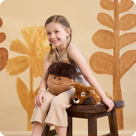 Young girl sitting on a stool with Laying Down Deer Warmies® against a floral-patterned wall.