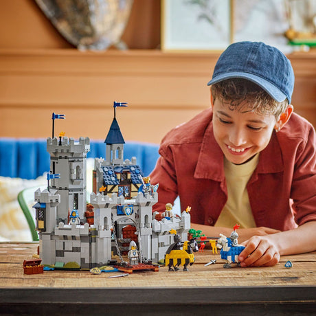 A young boy wearing a blue cap and red jacket joyfully playing with a LEGO Creator: Medieval Horse Knight Castle, featuring a detailed gray castle, knights, and horses.