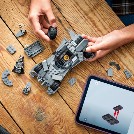 Person assembling a LEGO Super Heroes DC: Batman v Superman™ Batmobile™ model on a wooden table with a tablet displaying building instructions.