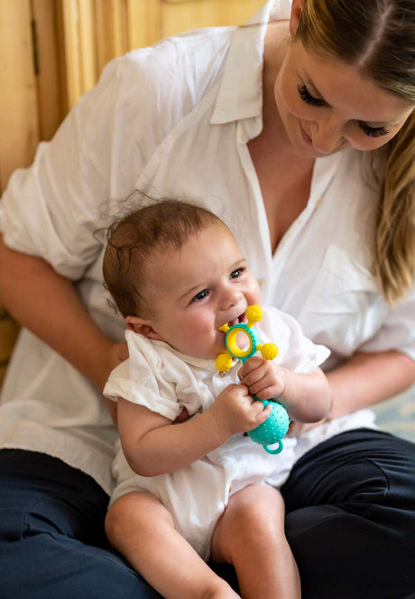 Woman holding a baby who is holding Gumlii Teether Rattle, all wearing white shirts.