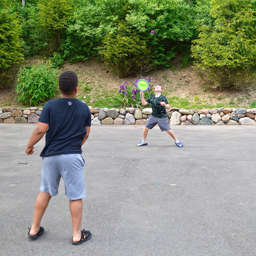 Two people playing with a Koosh® Paddle Play Set in an outdoor setting with greenery.