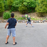 Two people playing with a Koosh® Paddle Play Set in an outdoor setting with greenery.