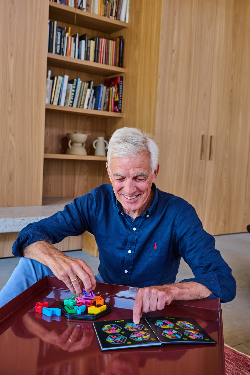 Man playing with colorful IQ Deluxe - Hexagon puzzle pieces on a table in a room with bookshelves.
