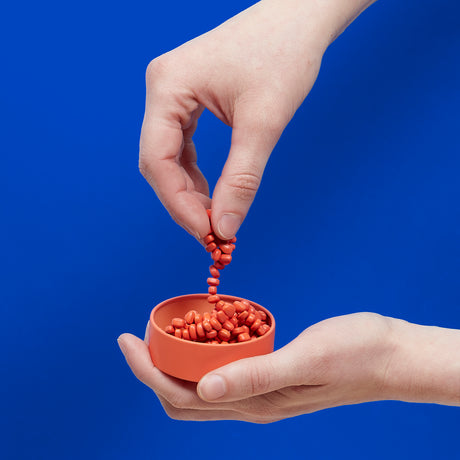 Hand pouring Mini Matte Crags Coral a small orange tin against a blue background