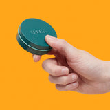 Hand holding a teal Mini Matte Crags Jade container against an orange background