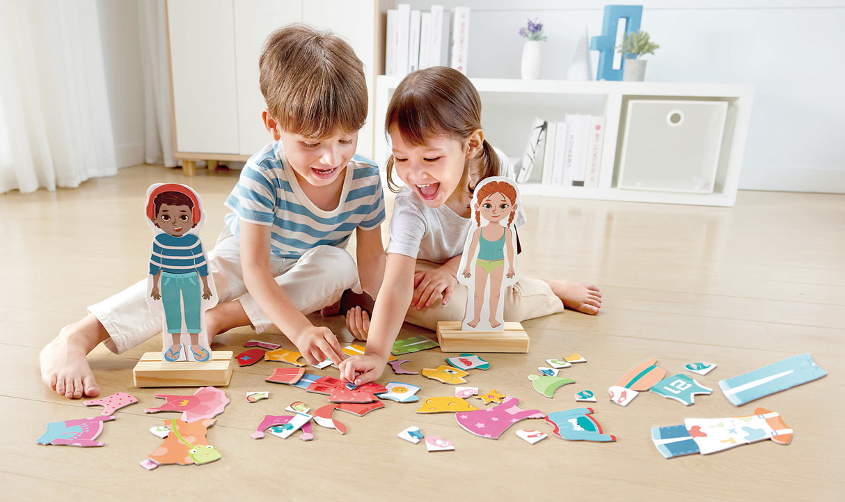 Two children playing with Dress-Up Magnetic Puzzle wooden dolls and colorful pieces on a light-colored floor.