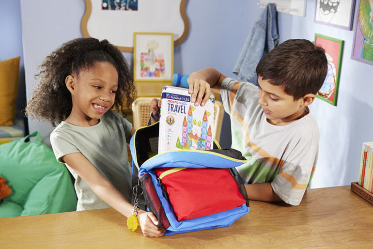 Two children looking at a Magna-Tiles microMags Travel Deluxe 55 Piece Set in a colorful bag on a table.