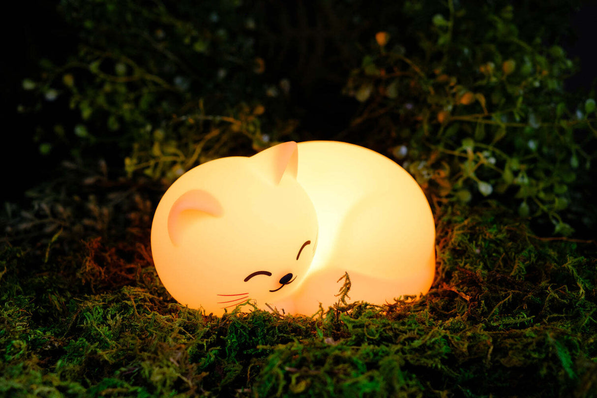 World of Mushi Ambient Squishy Cat Light night light glowing with a warm yellow light, nestled in green moss outdoors at night.