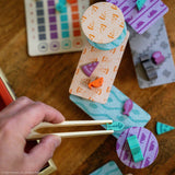 A close-up of a hand using wooden tweezers to manipulate small colorful game pieces on a printed board, with a visible hand and real tabletop environment making this a Lifestyle image rather than Product/Packaging from the Cats Knocking Things Off Ledges Balance Game.