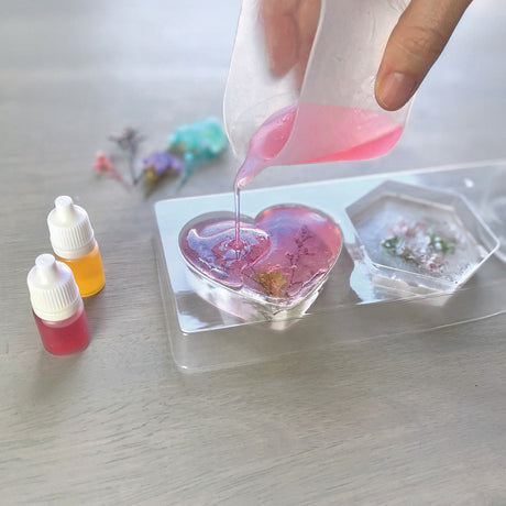 Person pouring soap into a heart-shaped mold on a light wooden surface.