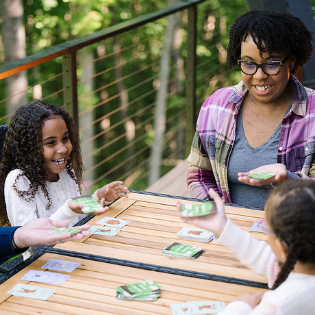 Three people playing Moose Match Mayhem Card Game outdoors on a wooden table with greenery in the background.