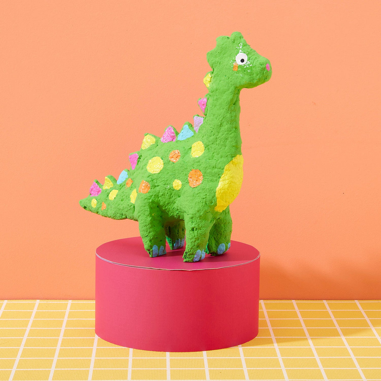 Green dinosaur figurine with colorful spots on a pink stand against an orange background made with the Paper Mache All Day! kit