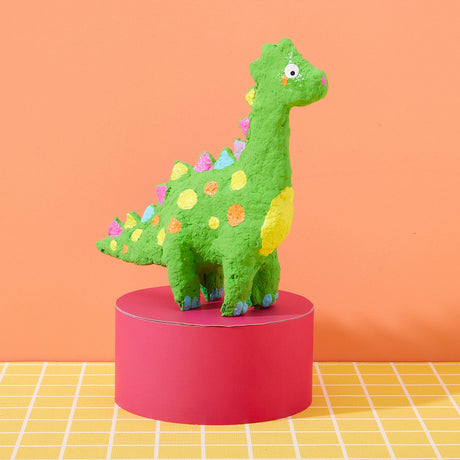 Green dinosaur figurine with colorful spots on a pink stand against an orange background made with the Paper Mache All Day! kit