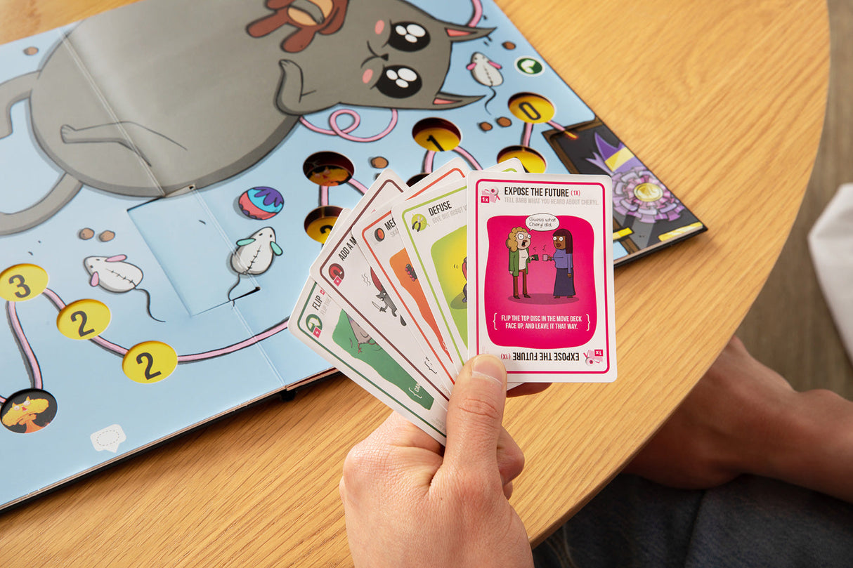 Person holding playing cards from the Exploding Kittens The Board Game on a wooden table