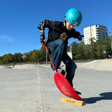 Child on a The Spooner Pro Born to Ride Training System - 26" wearing a blue helmet at a skate park.