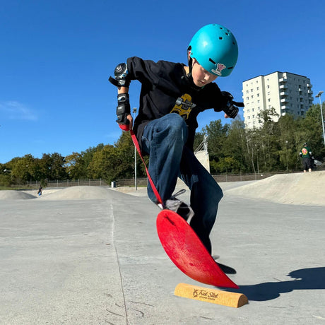 Child on a The Spooner Pro Born to Ride Training System - 26" wearing a blue helmet at a skate park.